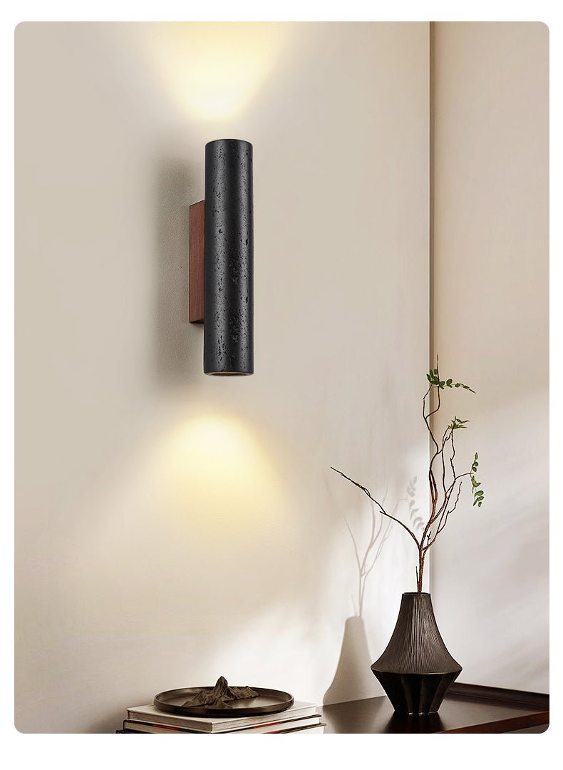 A Clérachic cylinder wall light lights a cream wall above a dark vase, a branch and a stack of books on a console.