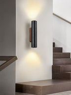 A Clérachic wall light shines up and down a pale stairwell wall, with dark wooden stairs rising to the right.