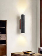 A Clérachic black cylinder wall light glows above a timber console holding two shallow bowls, with a wooden cabinet beside.