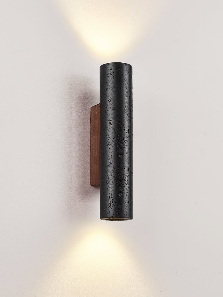 A Clérachic black travertine cylinder wall light with up and down beams against a soft pinkish-grey wall.