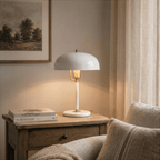 Clérachic white dome table lamp with brass fittings lit on a wooden nightstand beside stacked books and a linen curtain.