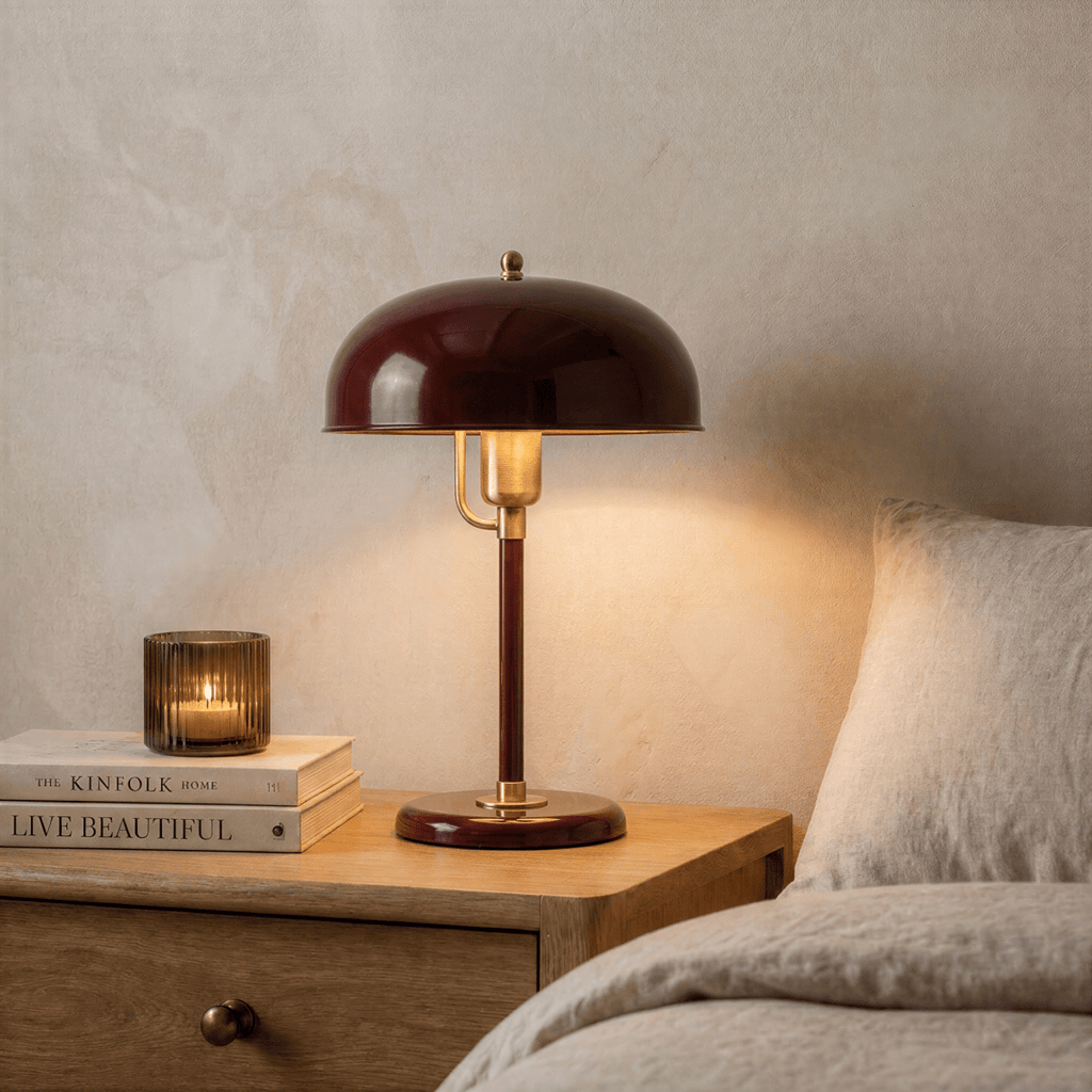 Clérachic red dome table lamp with brass fittings lit on a wooden nightstand beside stacked books and a glass candle holder.