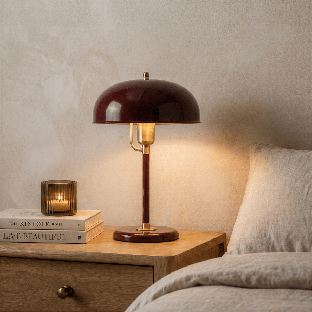 Clérachic red dome table lamp with brass fittings lit on a wooden nightstand beside stacked books and a glass candle holder.