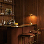 A lit Clérachic gold table lamp glows on a brass bar counter lined with bottles and a bowl of lemons.