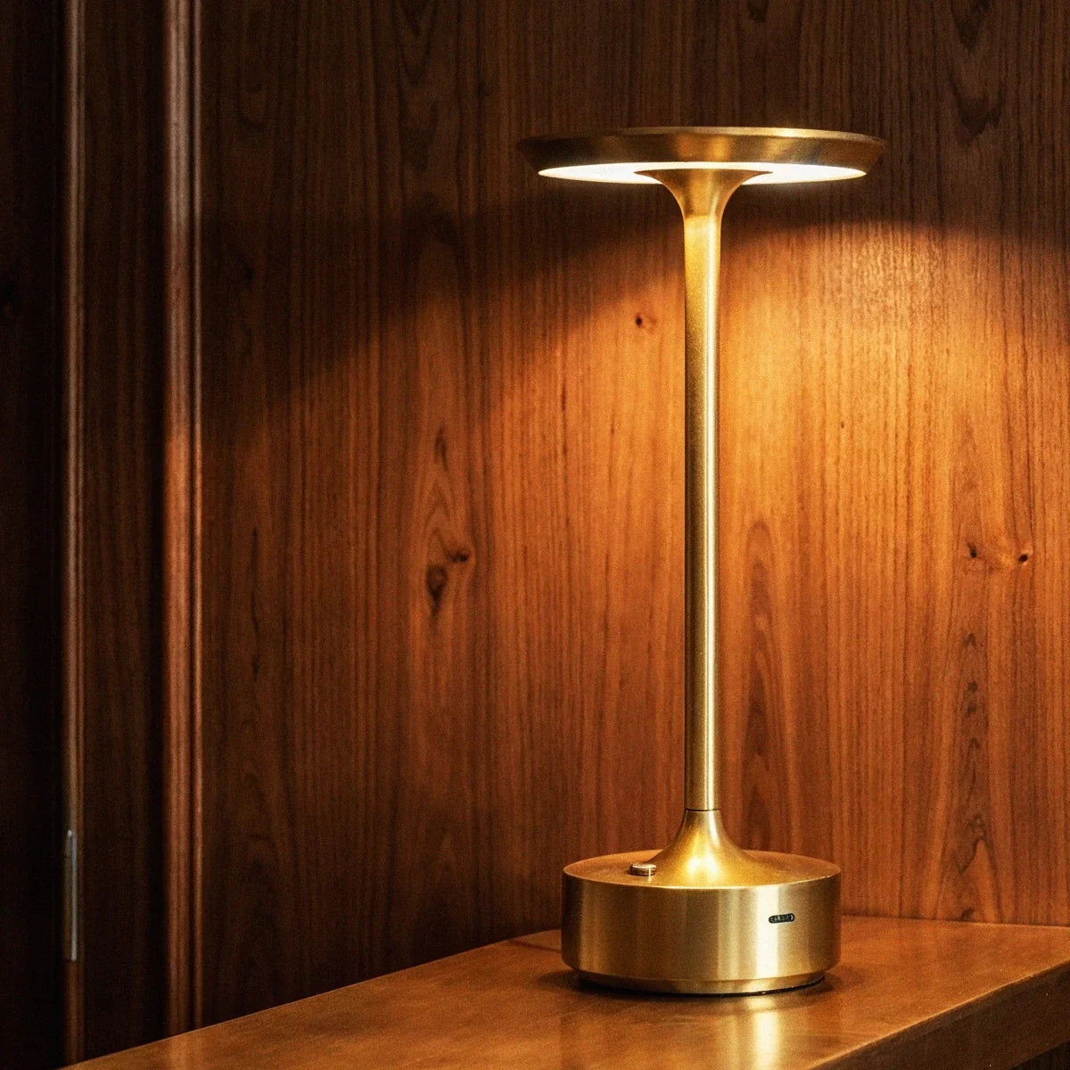 Clérachic's gold cordless lamp glows warm against walnut wall panelling, lighting a wood-topped side table.