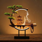 Clérachic incense-burner lamp lit warm on a wood table, a standing white figure and pagoda, beads hanging low.
