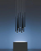 Unlit-looking cluster of black needle-thin pendants glowing at the tips, hung above a dark block against a blue wall, a Clérachic pendant.