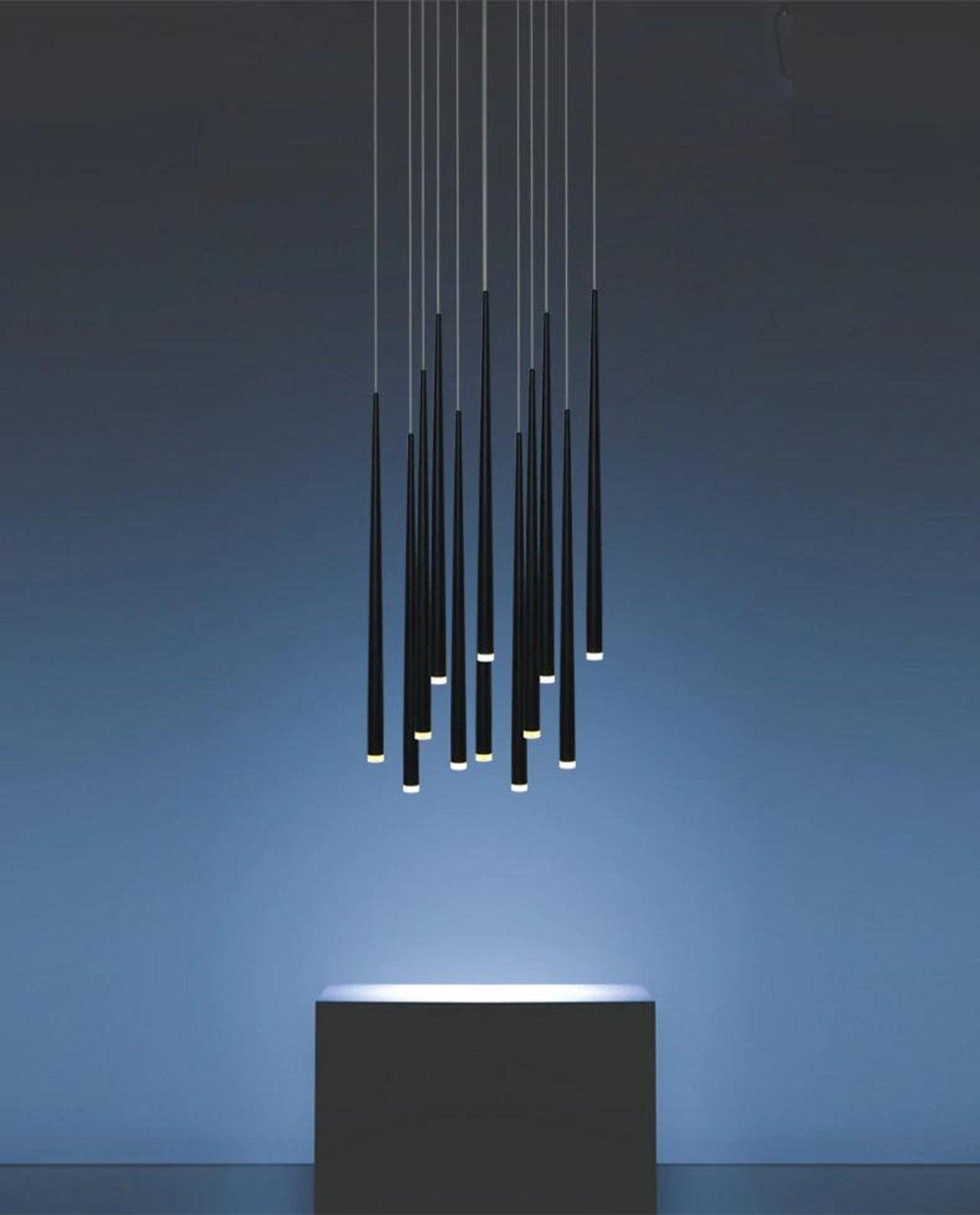 Unlit-looking cluster of black needle-thin pendants glowing at the tips, hung above a dark block against a blue wall, a Clérachic pendant.
