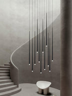 Tall group of black tapered pendants dropping beside a curving plaster staircase, with a pale sculptural stool below, this Clérachic pendant.