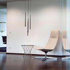 Three slim black tapered pendants glow at their tips beside a pale lounge chair on a dark wooden floor, a Clérachic pendant.