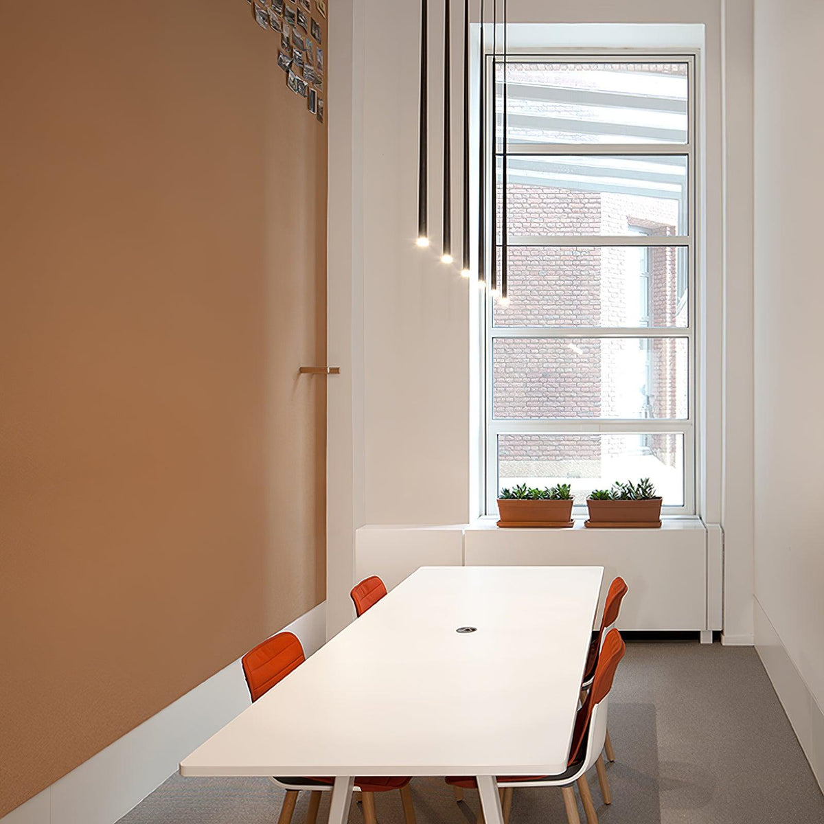 A row of slim black pendants steps down alongside a window above a long white table with orange chairs, from Clérachic.