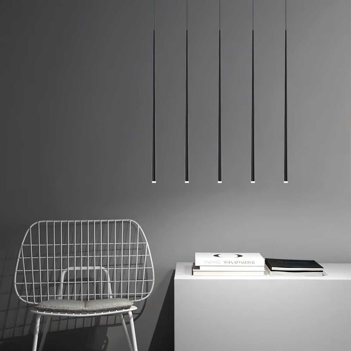 Five very long black pendant tubes hang evenly over a white cabinet and a wire chair on a grey wall, from Clérachic.