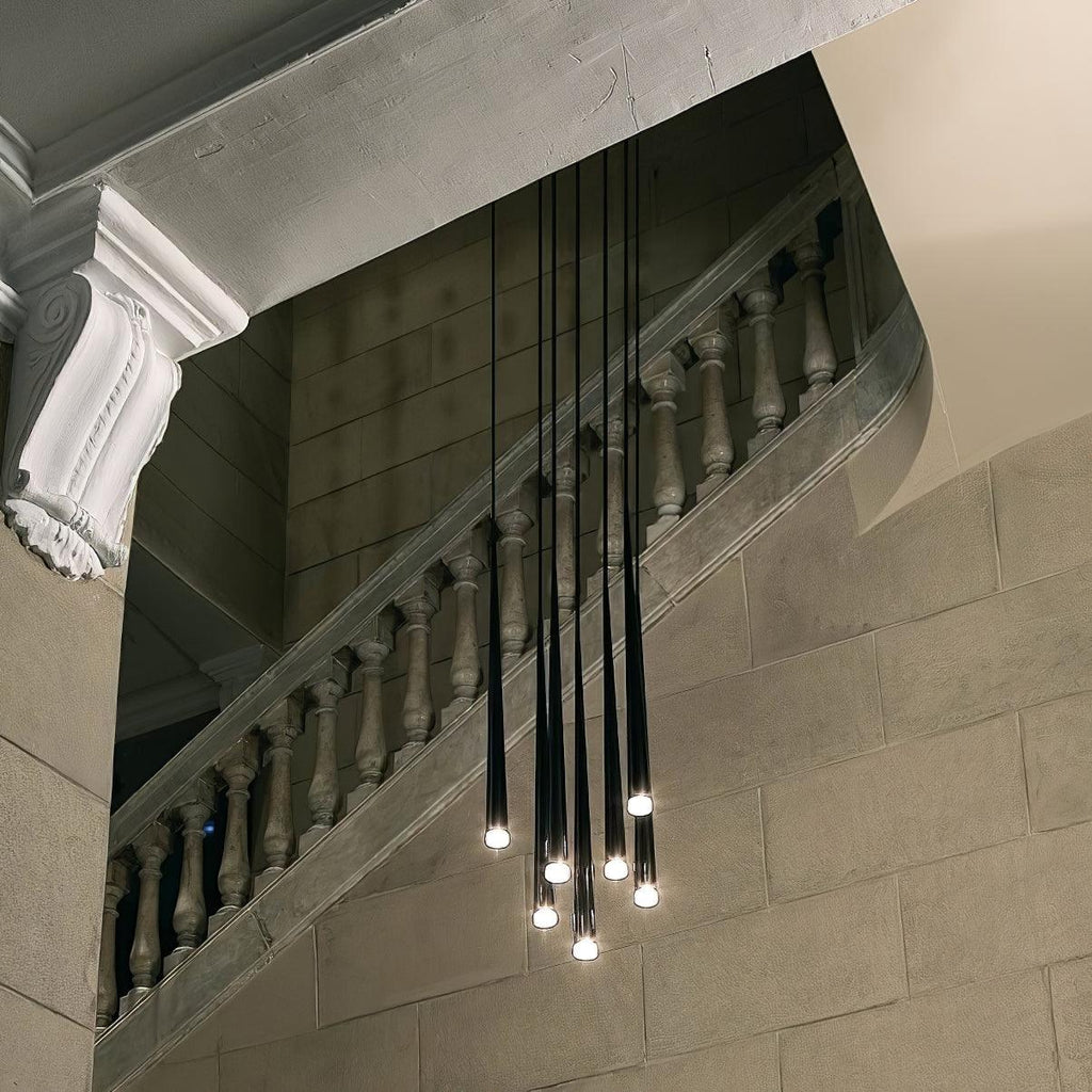 Black tapered pendants hang at varied heights in front of a stone balustrade staircase, each tip glowing, a Clérachic pendant.