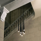 Black tapered pendants hang at varied heights in front of a stone balustrade staircase, each tip glowing, a Clérachic pendant.
