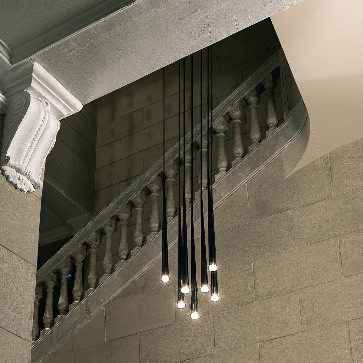 Black tapered pendants hang at varied heights in front of a stone balustrade staircase, each tip glowing, a Clérachic pendant.