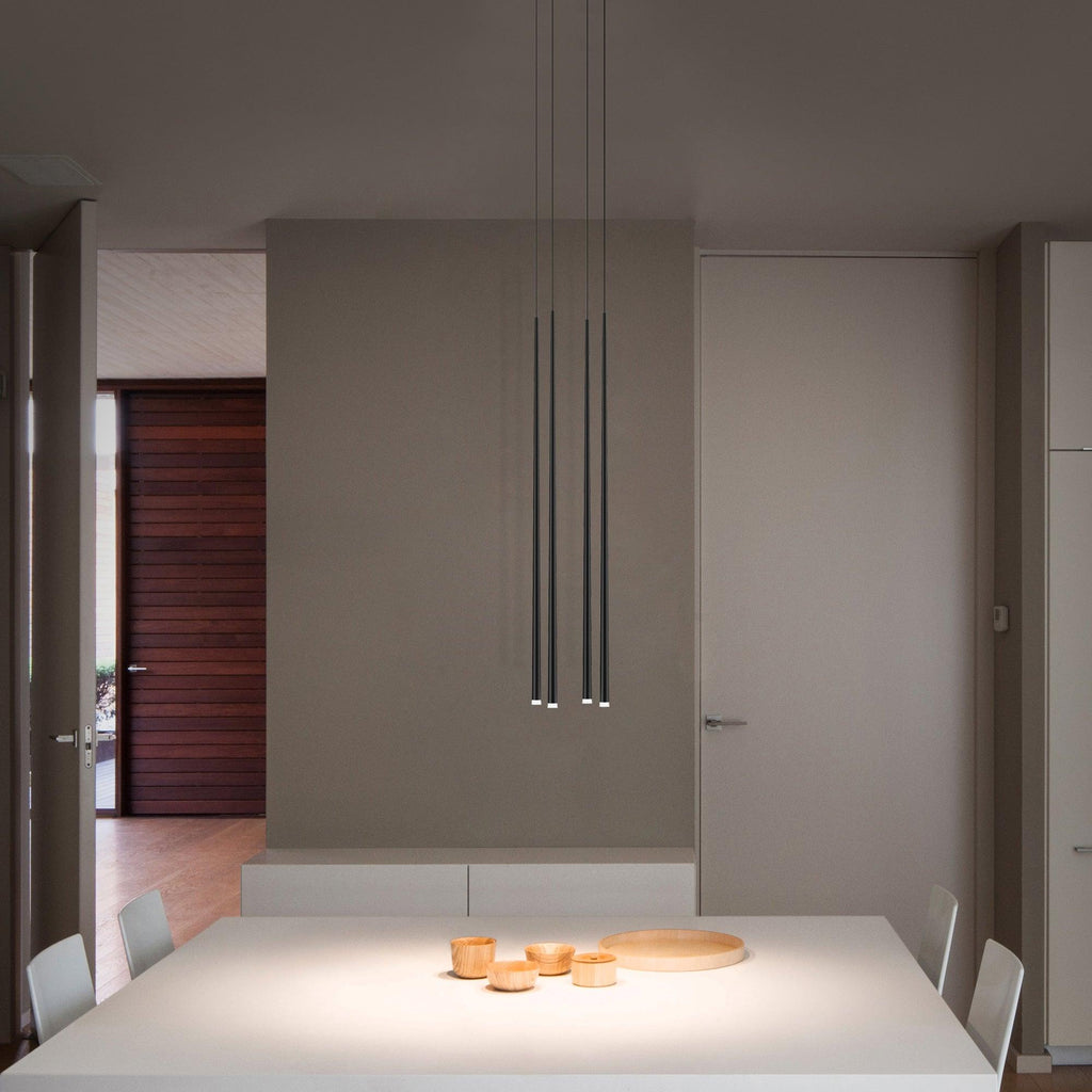 Three slim black pendants hang low over a white dining table with small wooden bowls in a calm taupe room, from Clérachic.