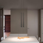 Three slim black pendants hang low over a white dining table with small wooden bowls in a calm taupe room, from Clérachic.