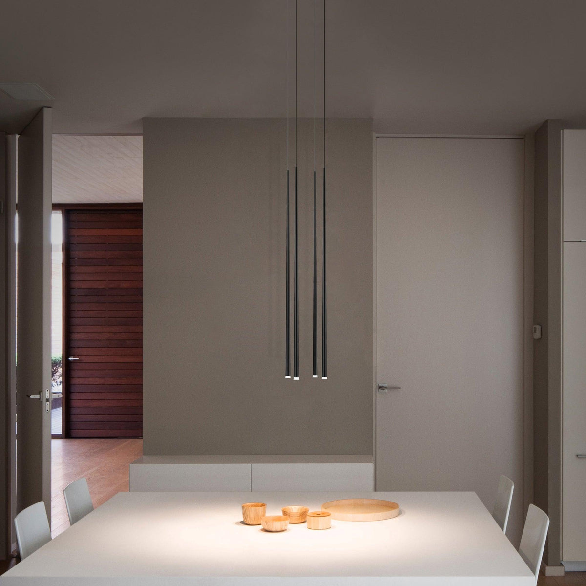 Three slim black pendants hang low over a white dining table with small wooden bowls in a calm taupe room, from Clérachic.