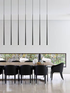 Seven long black tapered pendants in a line above a dark dining table with black chairs and a patterned screen, a Clérachic pendant.