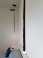 Single long black tube hangs close to the camera in a narrow room with a bed and blue cushion, a Clérachic pendant.