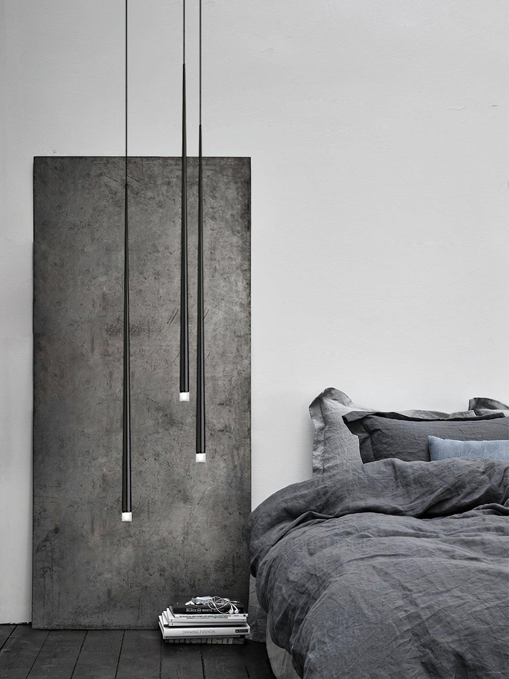 Three slim black pendants at different lengths hang beside a rough concrete panel and a bed with grey linen, from Clérachic.