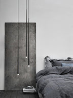 Three slim black pendants at different lengths hang beside a rough concrete panel and a bed with grey linen, from Clérachic.