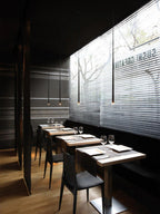 Slim black pendants with warm tips hang in a dark restaurant above small wooden tables and black chairs, from Clérachic.