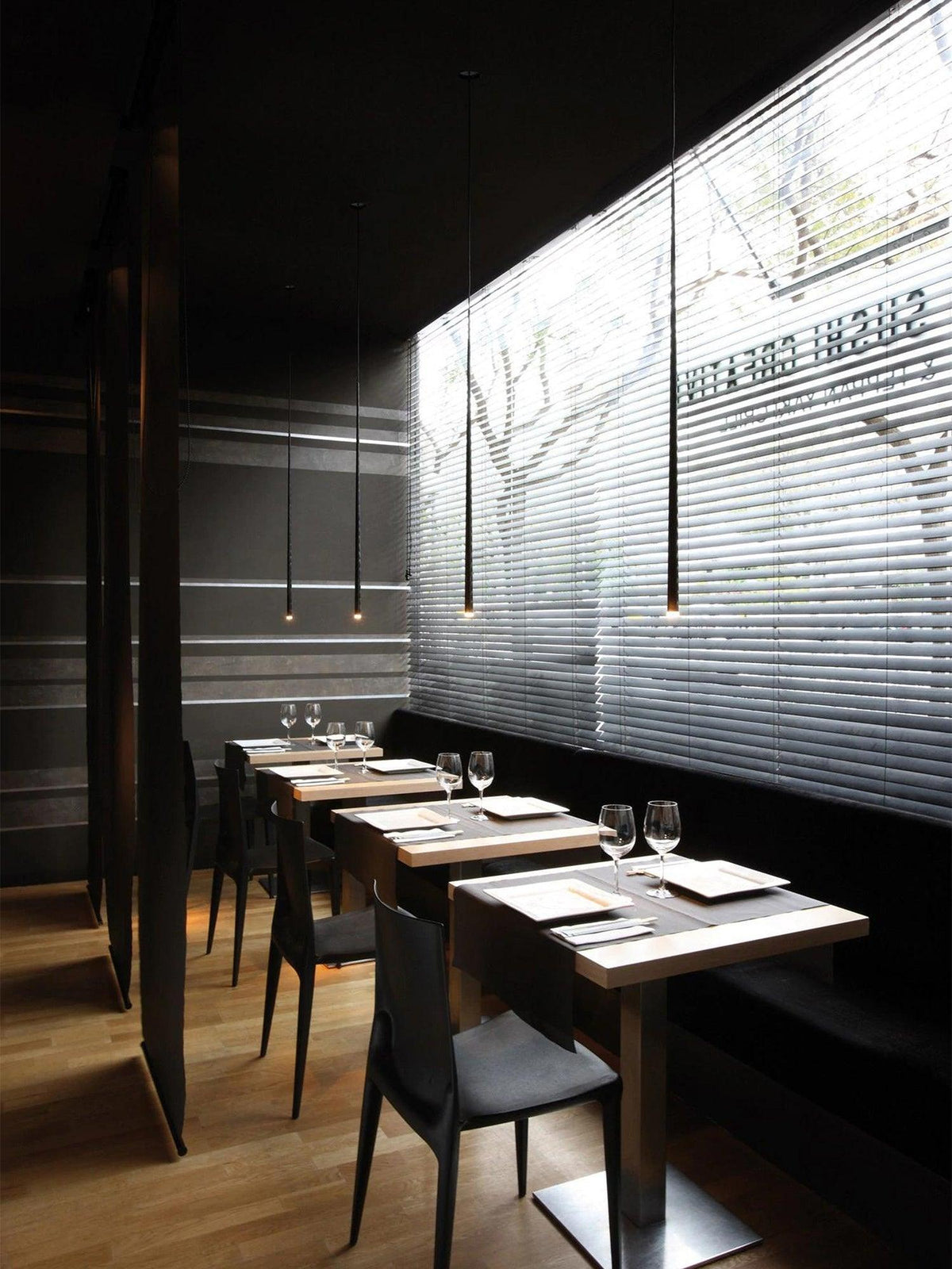 Slim black pendants with warm tips hang in a dark restaurant above small wooden tables and black chairs, from Clérachic.