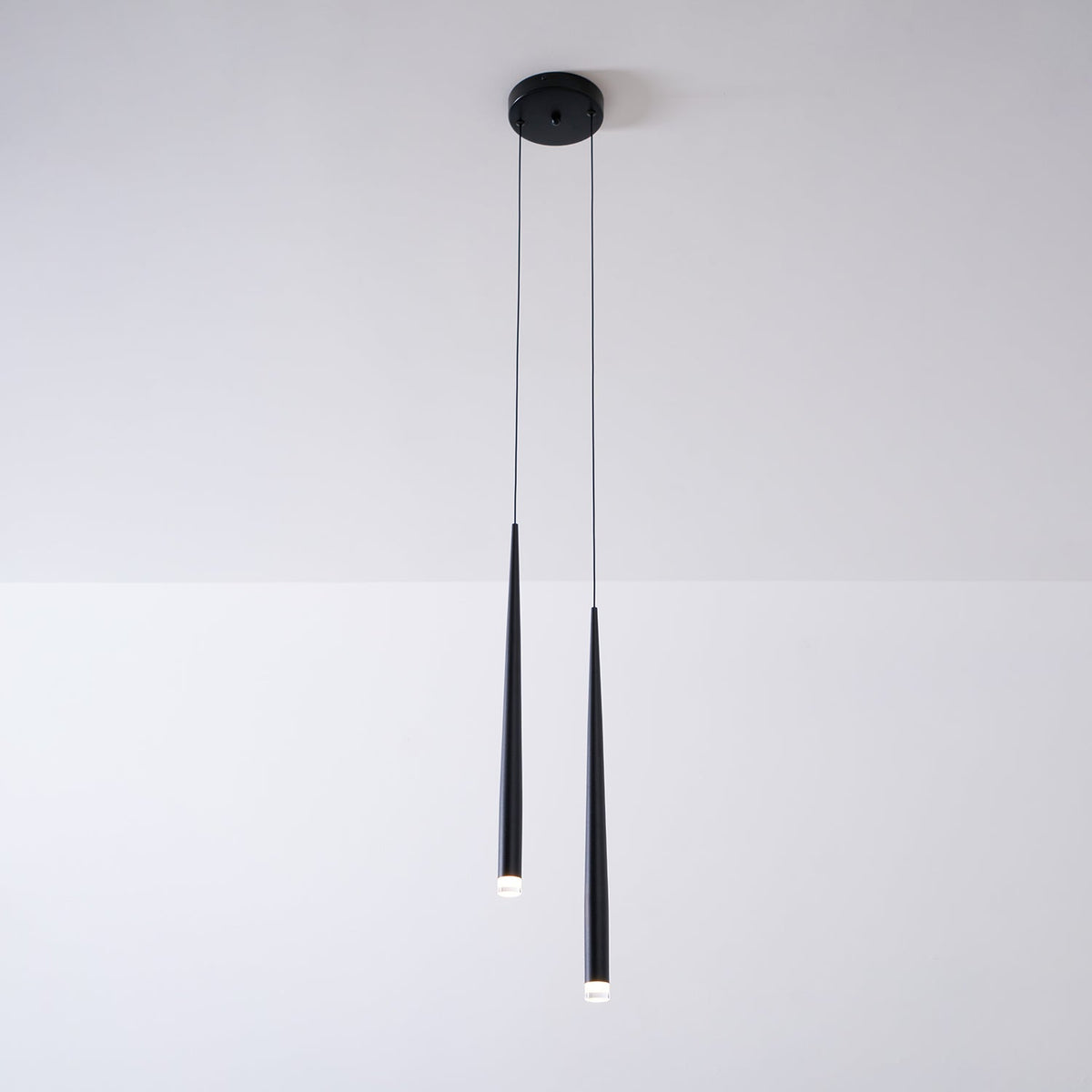 Two black tapered pendants from a round black ceiling canopy hang against a pale wall, their tips lit, from Clérachic.