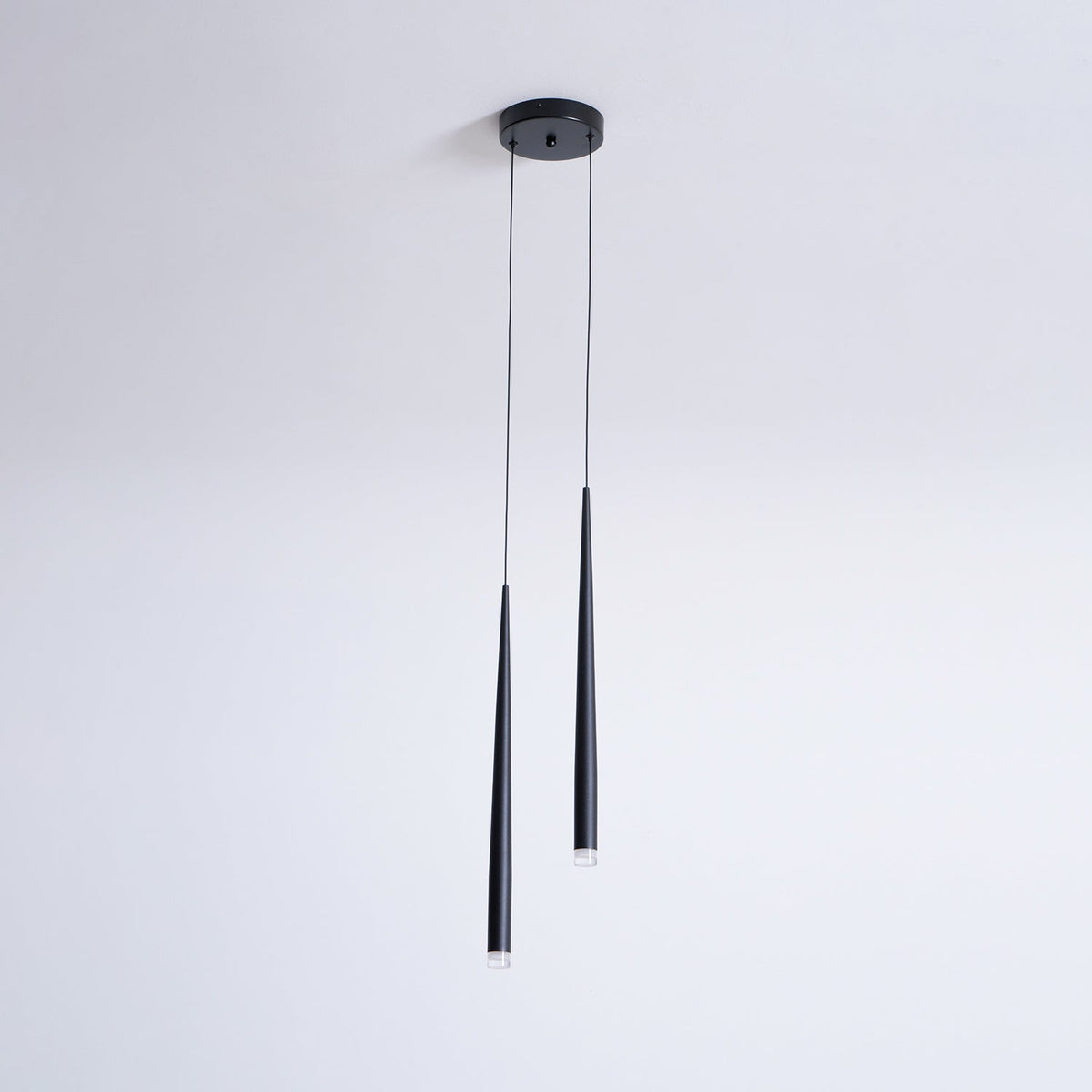 Two black tapered pendants of unequal length hang from a round canopy on a plain pale wall, from Clérachic.
