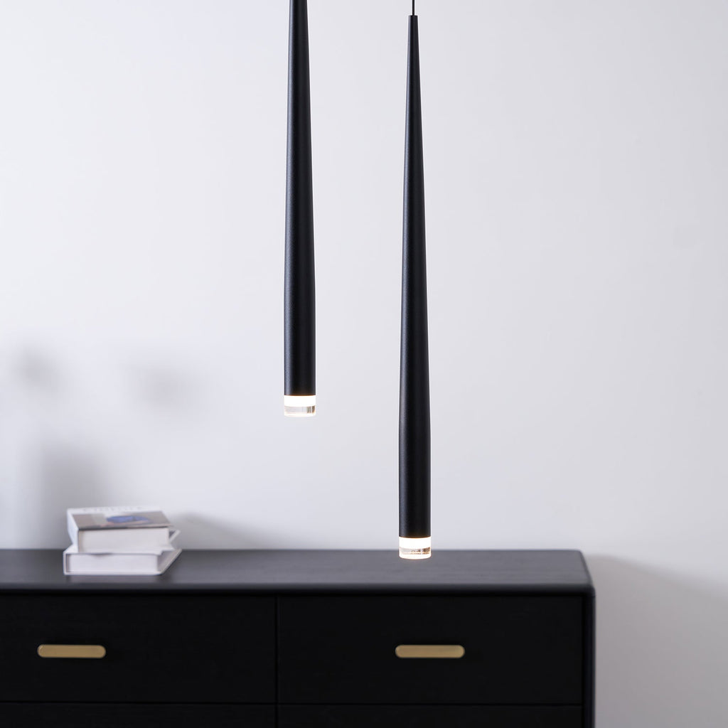 Two black tapered pendants close to the camera above a black chest with brass handles and a stack of books, from Clérachic.