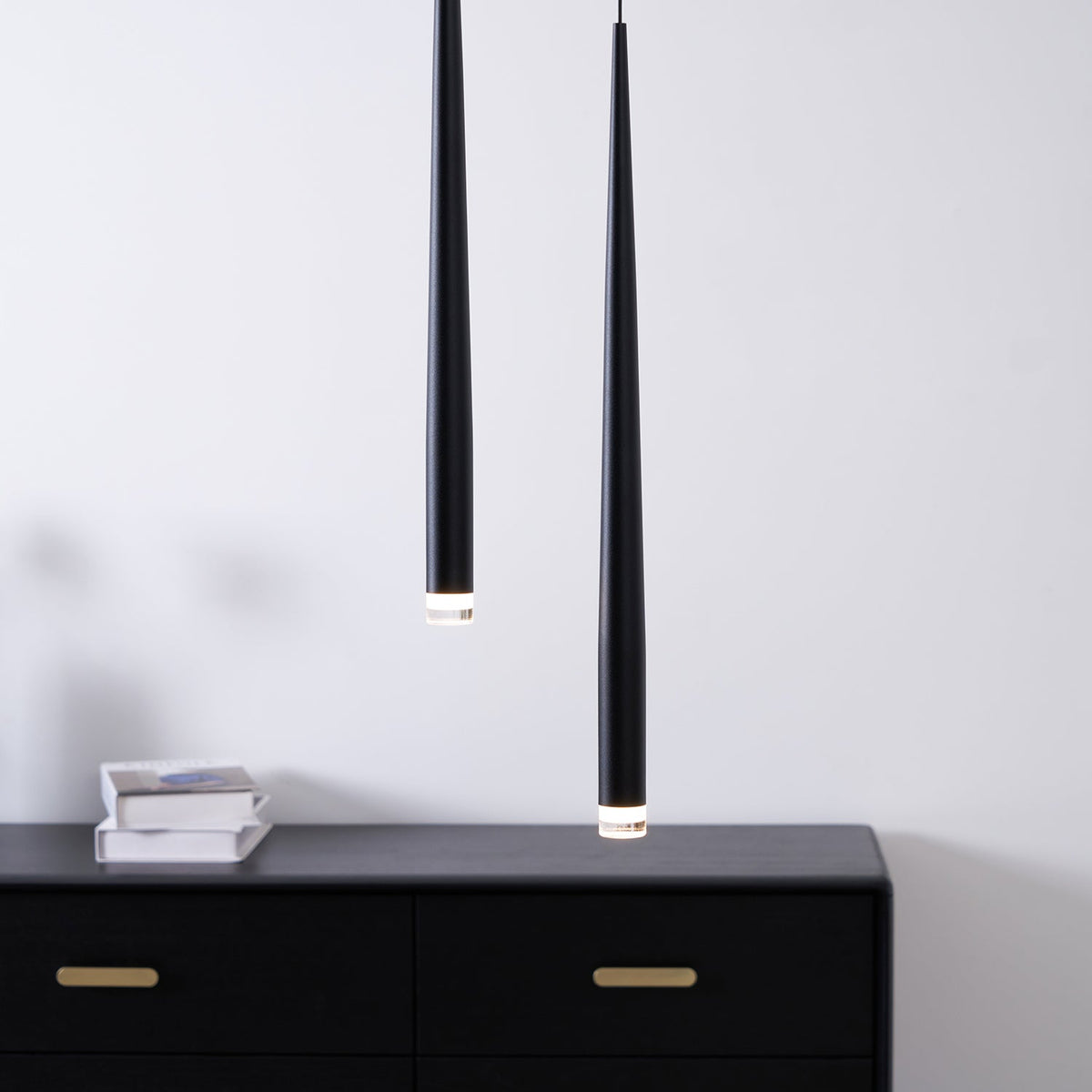Two black tapered pendants close to the camera above a black chest with brass handles and a stack of books, from Clérachic.