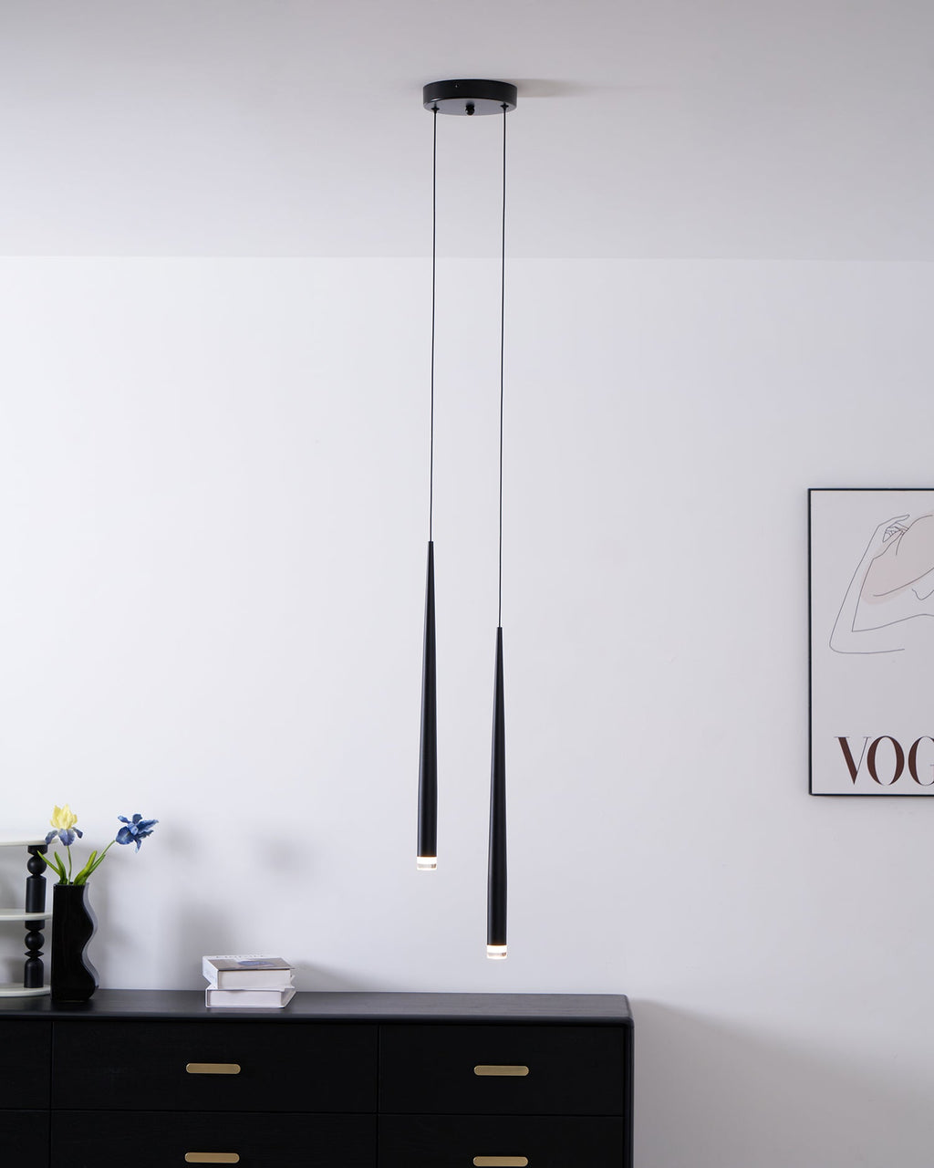 Two black tapered pendants hang above a black chest, with a vase of flowers and a framed print on the wall, a Clérachic pendant.