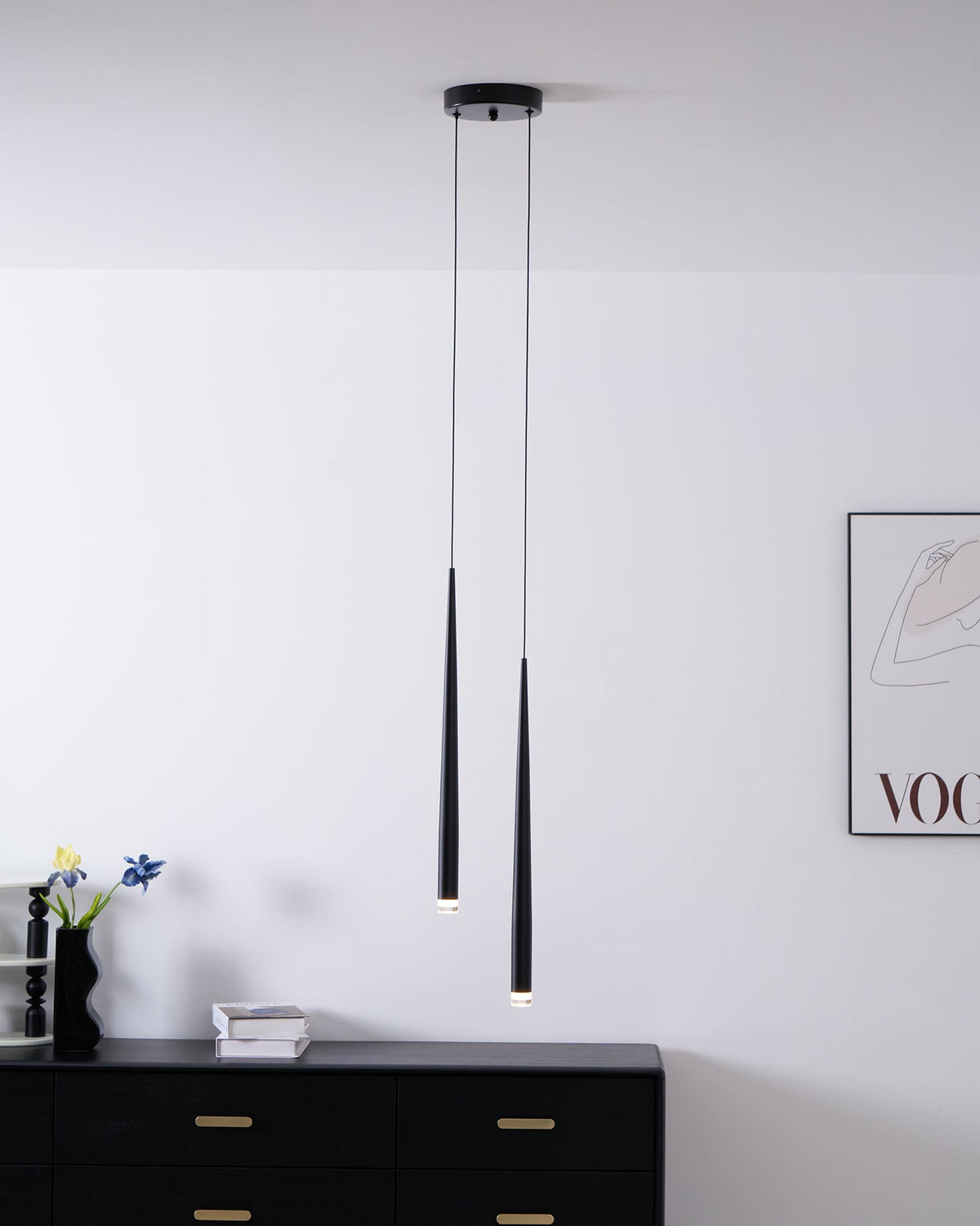 Two black tapered pendants hang above a black chest, with a vase of flowers and a framed print on the wall, a Clérachic pendant.