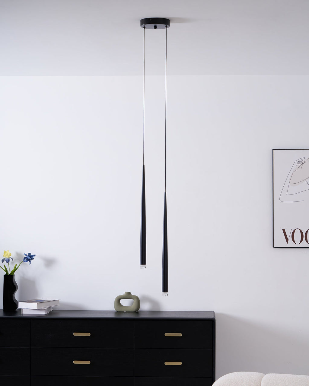 Wider room view of two black pendants above a black chest, with flowers, a vase and a cream sofa edge, a Clérachic pendant.