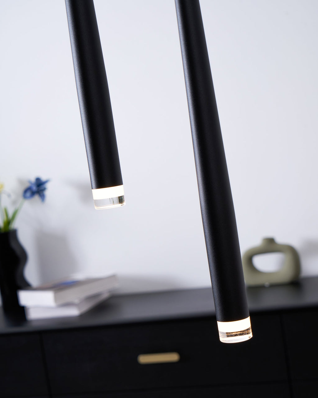 Angled close view of the lit lens ends of two black pendants above a black drawer chest with a brass pull, a Clérachic pendant.