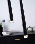 Angled close view of the lit lens ends of two black pendants above a black drawer chest with a brass pull, a Clérachic pendant.