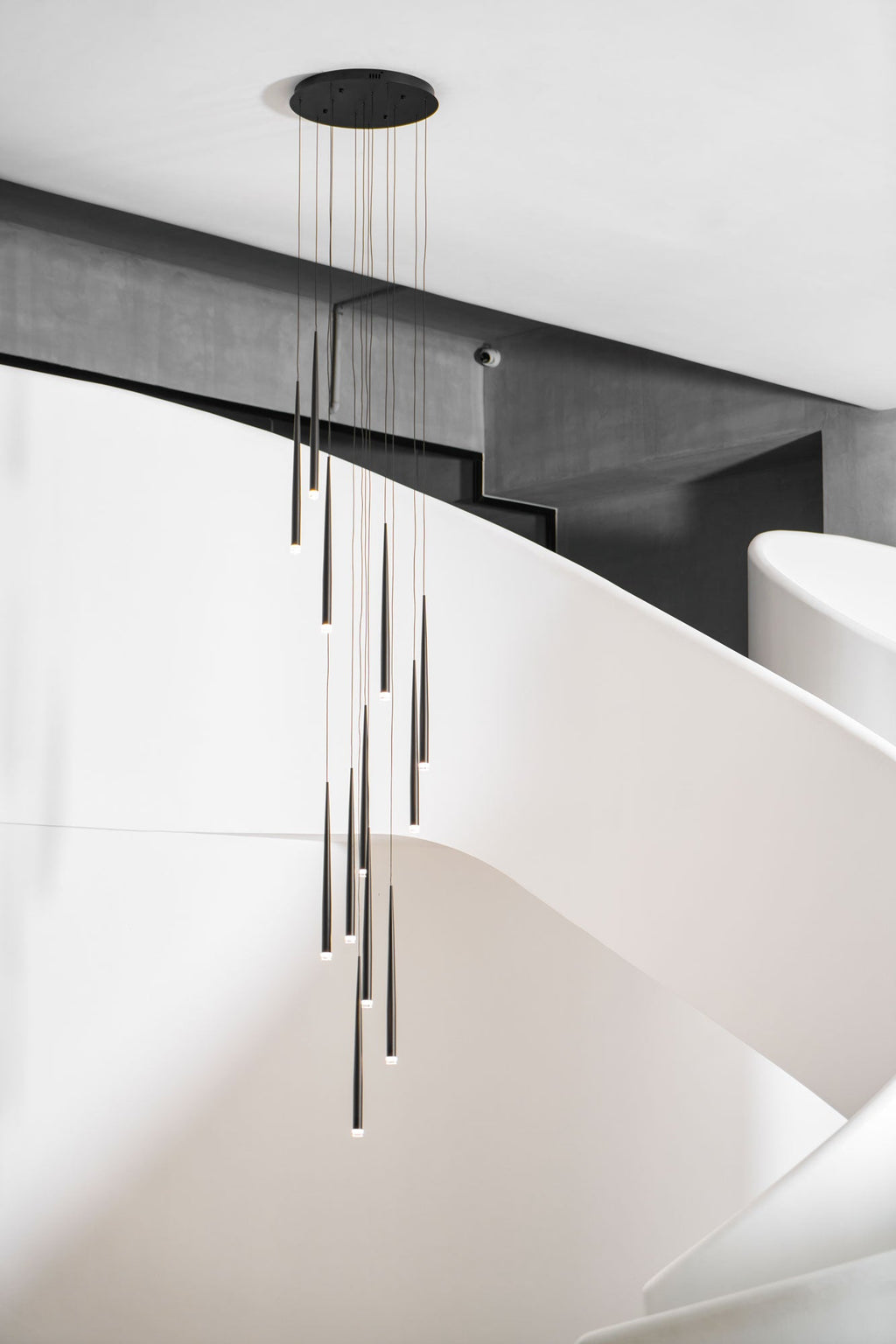 Round-canopy cluster of black tapered pendants hangs by a curving white staircase with a dark beam above, from Clérachic.
