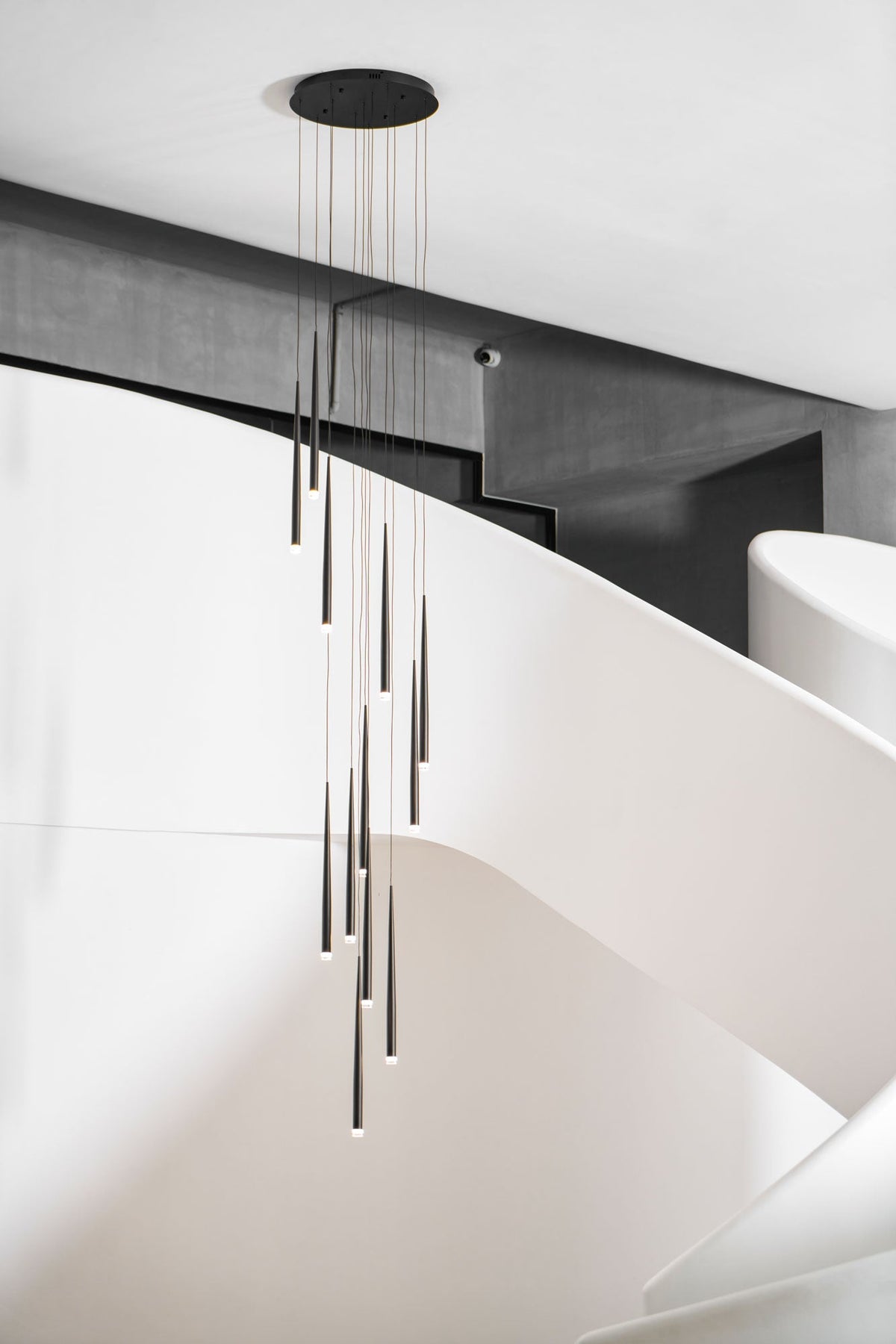 Round-canopy cluster of black tapered pendants hangs by a curving white staircase with a dark beam above, from Clérachic.