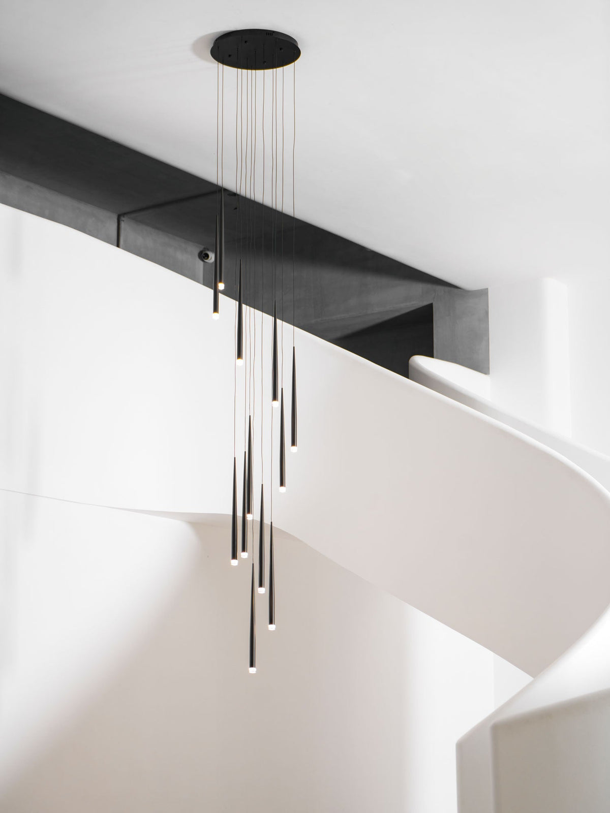 Black cone pendants of mixed length hang from a round canopy beside a white spiral staircase, a Clérachic pendant.