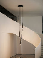 Lit black tapered pendants hang down the middle of a curved white stairwell, their tips glowing warm, from Clérachic.