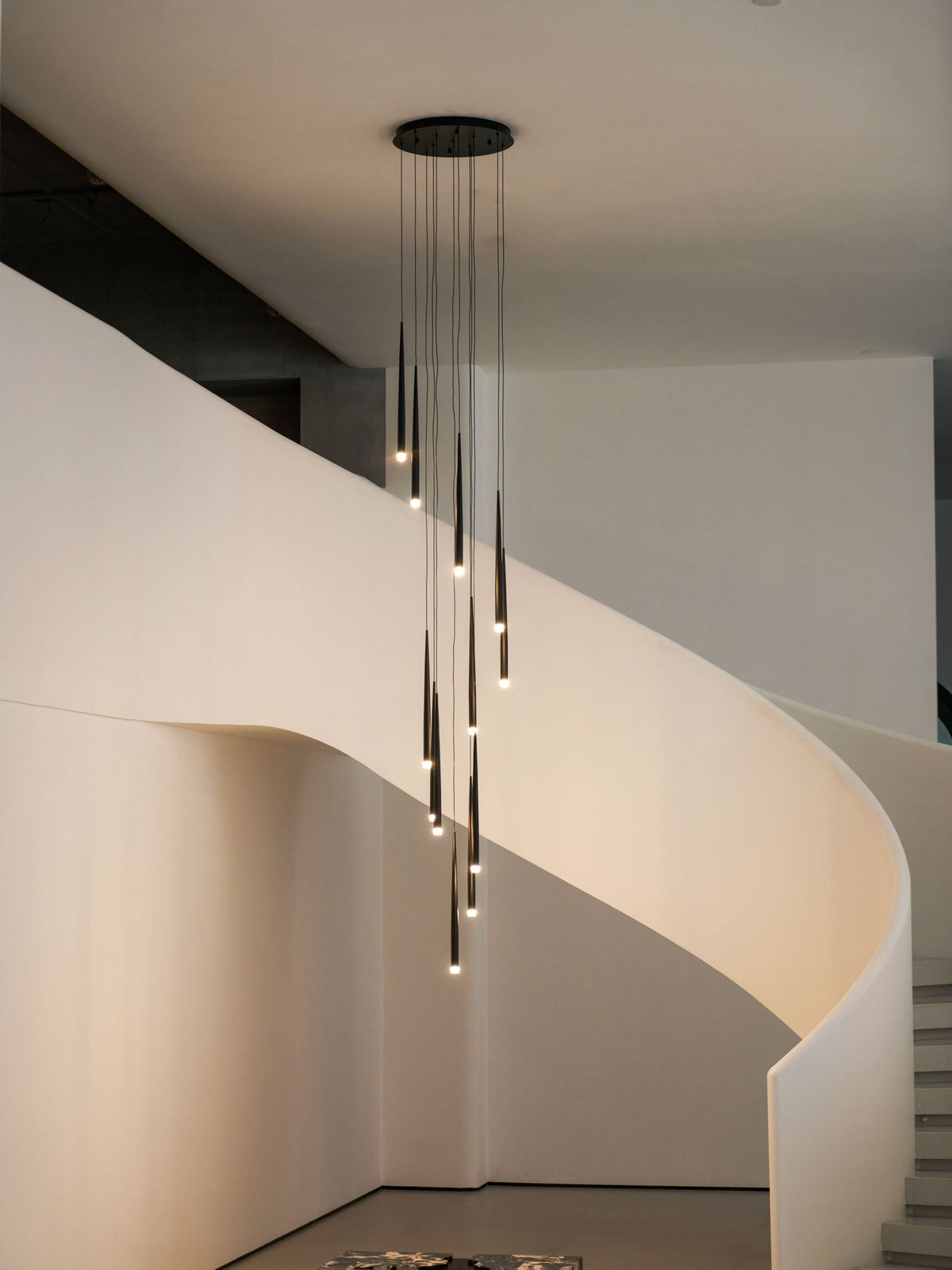 Lit black tapered pendants hang down the middle of a curved white stairwell, their tips glowing warm, from Clérachic.