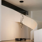 Wide view of a pendant cluster lit warm in a white stair hall, with a dark marble block on the floor, from Clérachic.