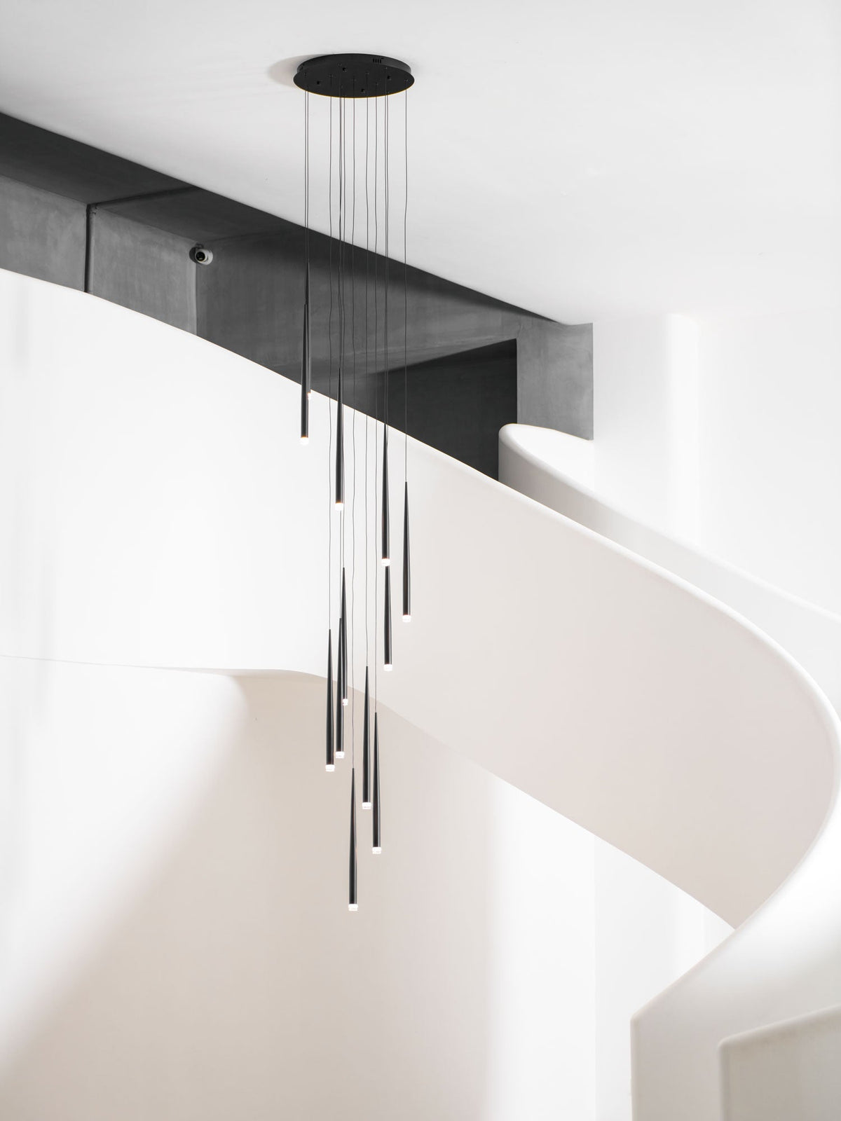 Black tapered pendants hang from a round canopy past a dark beam in a white spiral stairwell, from Clérachic.