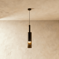 A black-tinted glass pendant light hangs on a slim rod, its cylindrical shade glowing warm against a plaster wall, Clérachic hero shot.