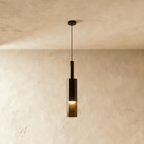 A black-tinted glass pendant light hangs on a slim rod, its cylindrical shade glowing warm against a plaster wall, Clérachic hero shot.