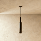 Unlit and straight-on, the black-tinted glass pendant's cylindrical shade hangs against a bare plaster wall, Clérachic hero shot.