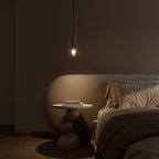 A single black-tinted pendant glows warm above a round nightstand beside a curved upholstered headboard, Clérachic lifestyle shot.