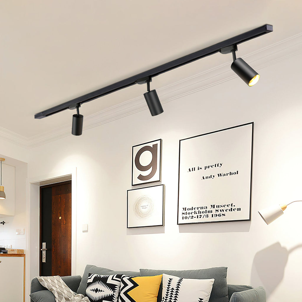 Clérachic black track with three black cylindrical heads above a living room wall with framed prints and a grey sofa.
