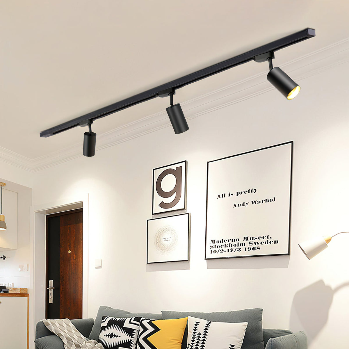 Clérachic black track with three black cylindrical heads above a living room wall with framed prints and a grey sofa.